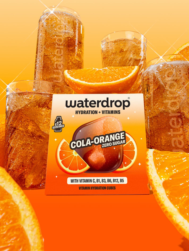Pudełko smaku Cola Orange z 12 szt. umieszczone pośrodku, w tle znajdują się szklanki waterdrop® wypełnione napojem, otoczone kawałkami pomarańczy.
