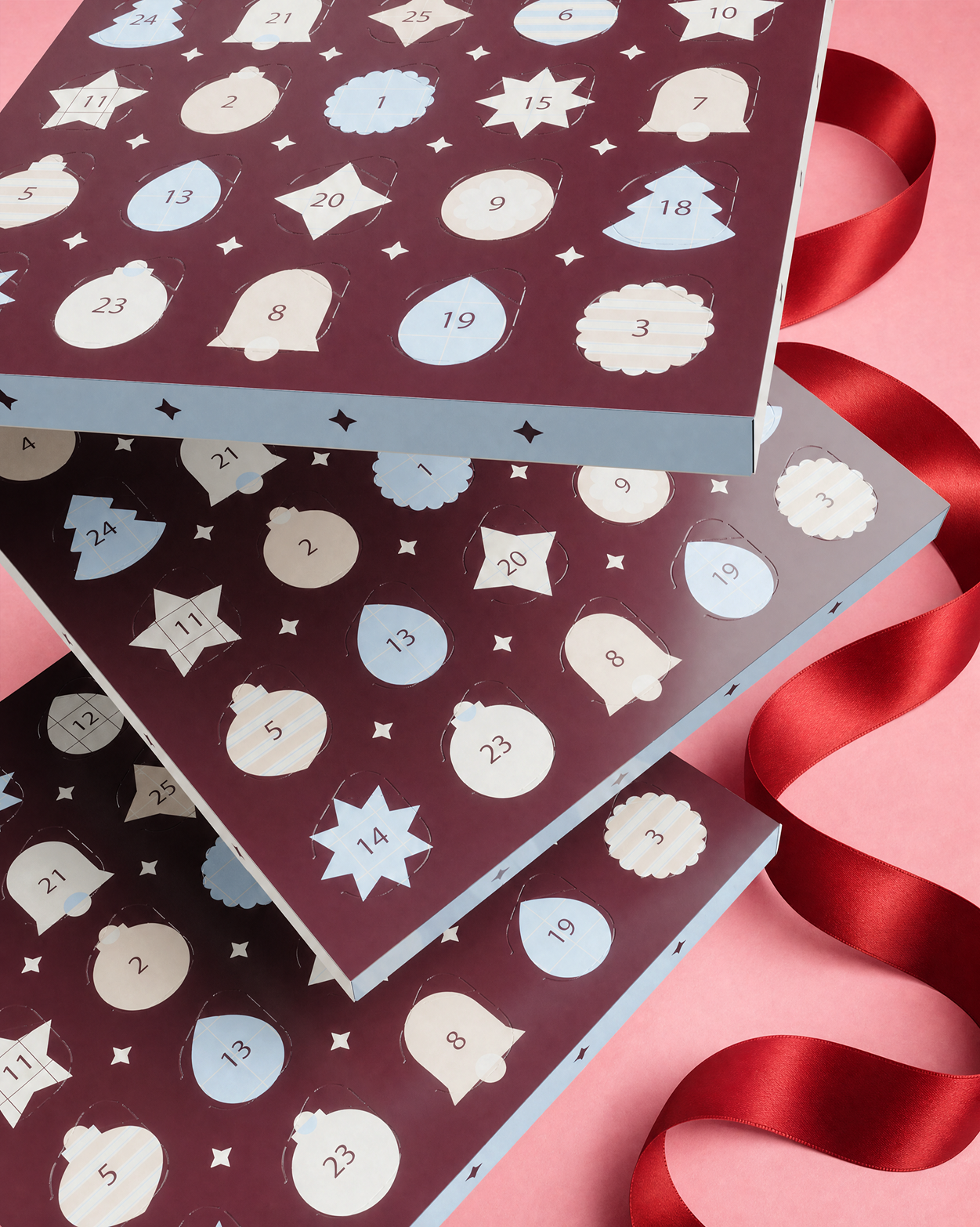 Trzy stojące Advent kalendarze z numerowanymi drzwiczkami i dekoracyjnymi kształtami, związane czerwonym wstążkiem na różowym tle.