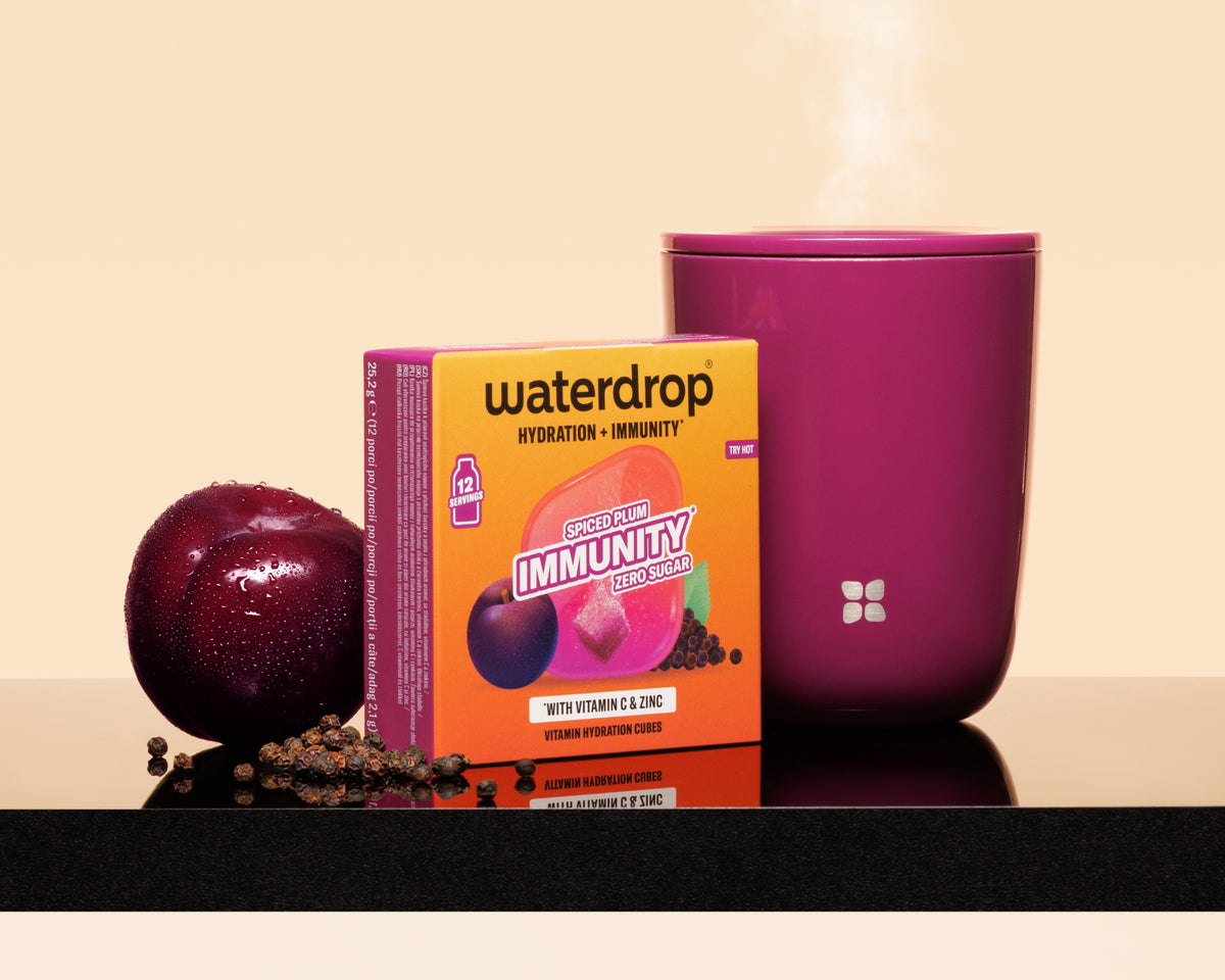 Purpurowa filiżanka z parą obok pomarańczowego pudełka Waterdrop, śliwka i rozsypane nasiona na błyszczącej powierzchni.